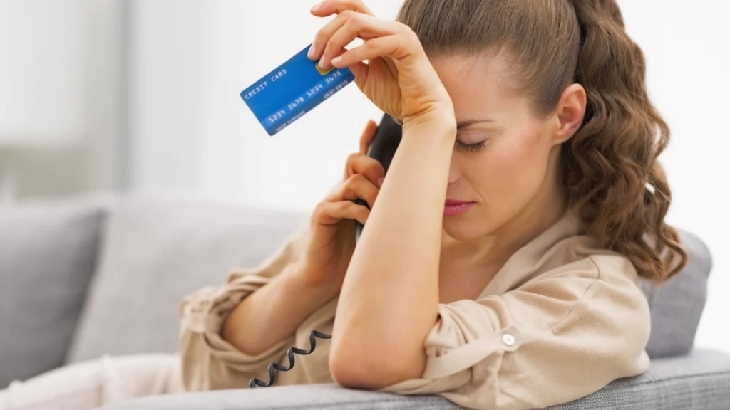 A woman looking stressed on the phone while holding her credit card, illustrating the anxiety of a **missed first credit card payment**.